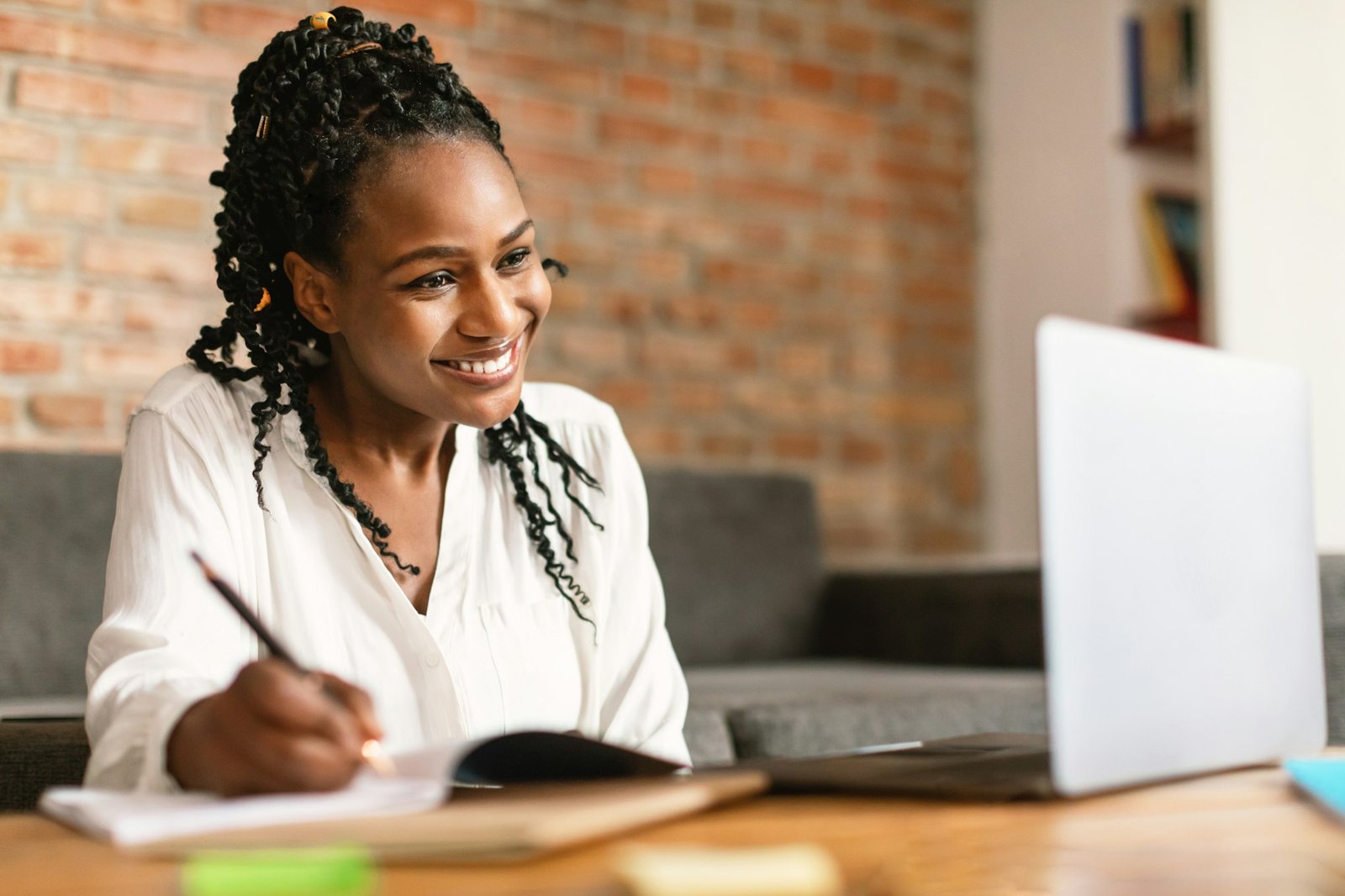 Excited black woman taking notes and using laptop, making video call while working or learning